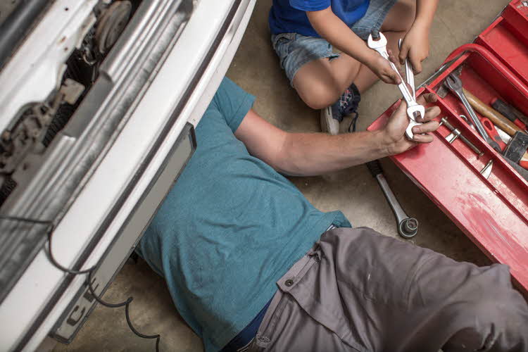 Fugleperspektiv på mann som ligger under bil og reparerer noe, barn gir ham verktøy. Gråtoner. Foto til artikkel om gaver til pappatyper på farsdag. 
