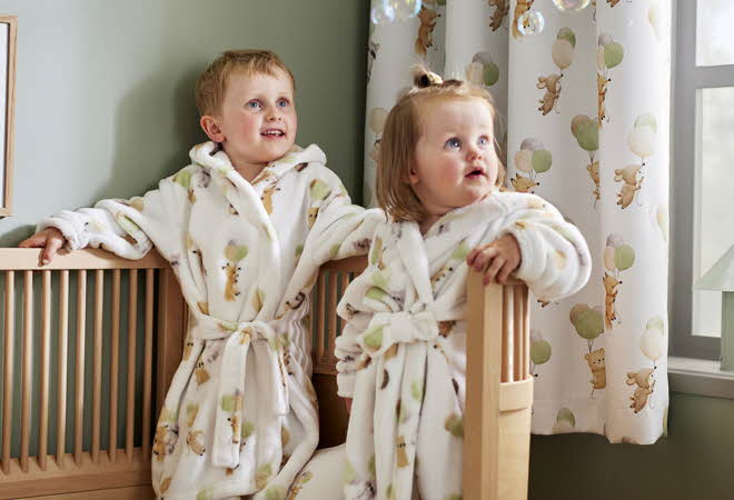 En liten gutt og jente står i like badekåper oppe i en seng