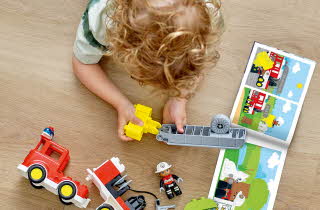 Barn bygger en LEGO Duplo brannbil på gulvet med instruksjonshefte ved siden av.