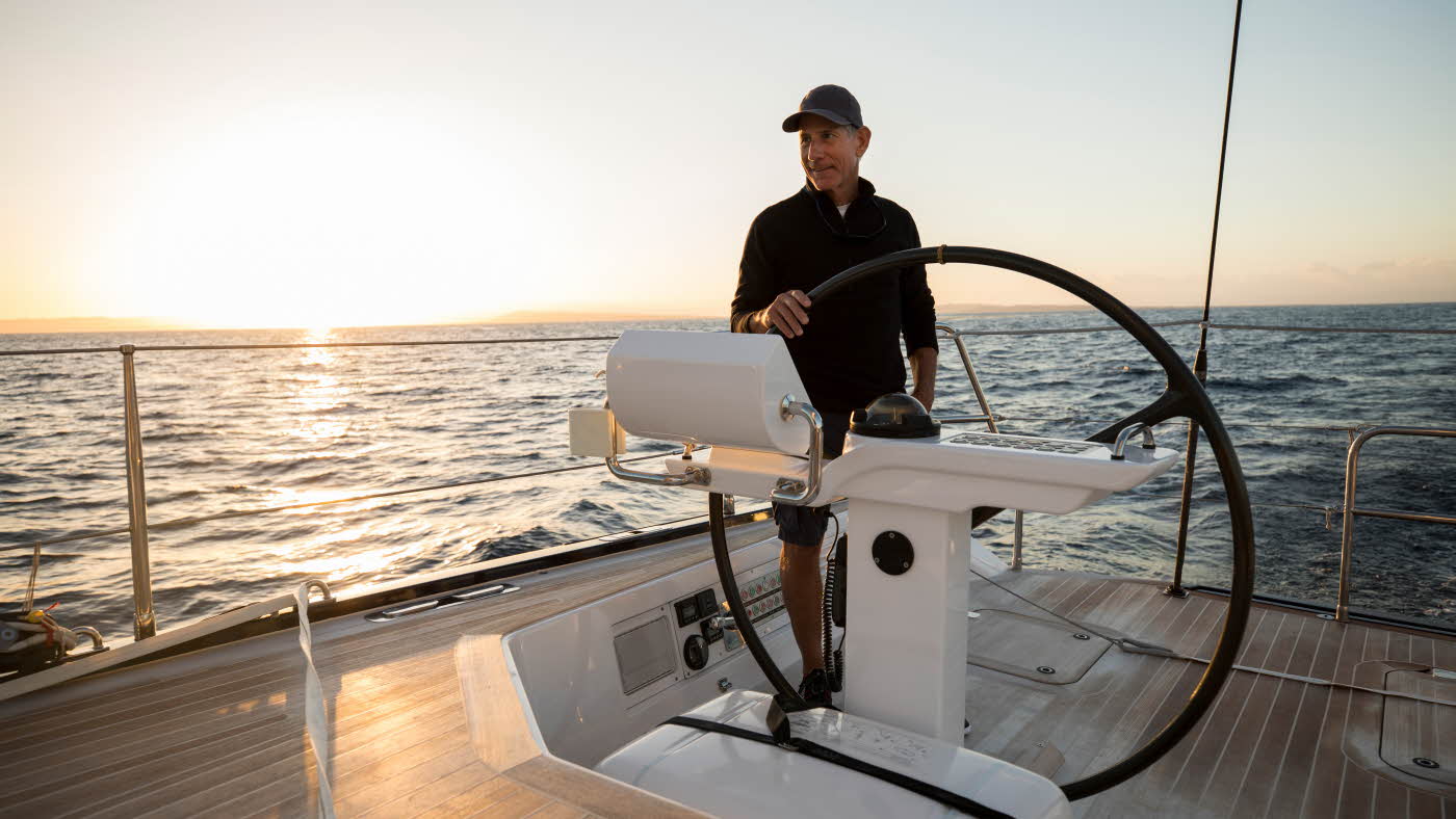 Mann med caps og mørk genser ved roret på en seilbåt på havet i solnedgangen