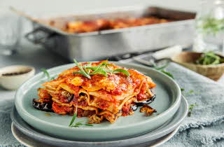 Et stykke lasagne på en blå tallerken, med mer lasagne i en form i bakgrunnen.