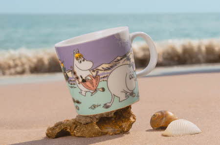 En Moomin Arabia kopp med motivet "Stranddag" står på et skjell på en strand
