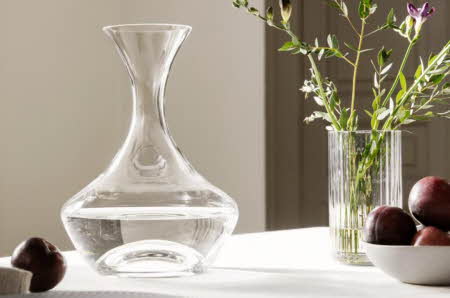 En karaffel står på et bord med en vase med blomster og en bolle med frukt.