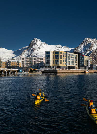 Personer padler i to kajakker i retning et havneområde med store, snødekte fjell og blå himmel i bakgrunnen.
