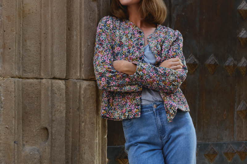 En kvinne lener seg på en gammel bygning, hun har på seg en blomstrete jakke og jeans