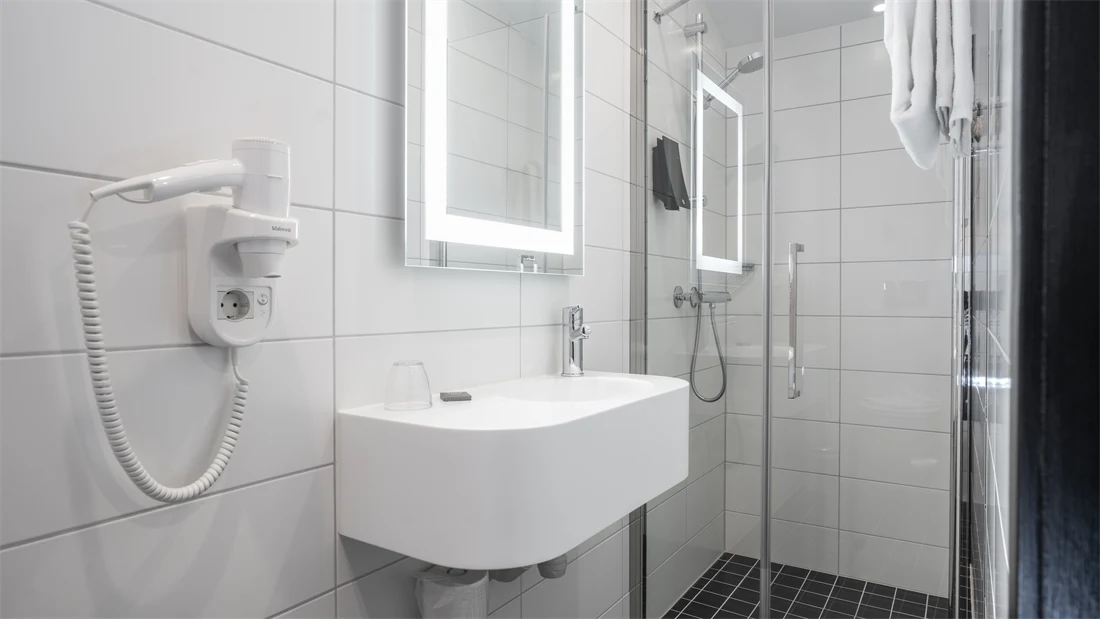 Veggfestet hårføn, servant, speil med innebygget lys, hvit kakel og dusj i baderom på Thon Hotel Astoria
