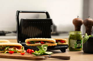 En sandwichgrill med to varme sandwicher, omgitt av ferske ingredienser på kjøkkenbenken.