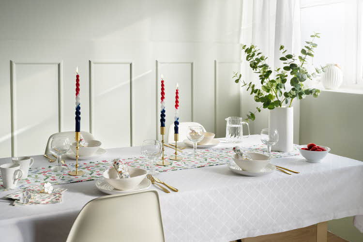 Bord dekket med hvit duk og mønstret løper, vase med grønne grener, hvitt servise, gullbestikk, vinglass, stearinlys i flaggets farger.