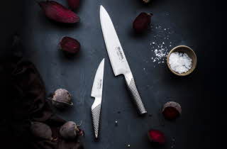 To kniver fra Global som ligger på et bord med grønnsaker rundt