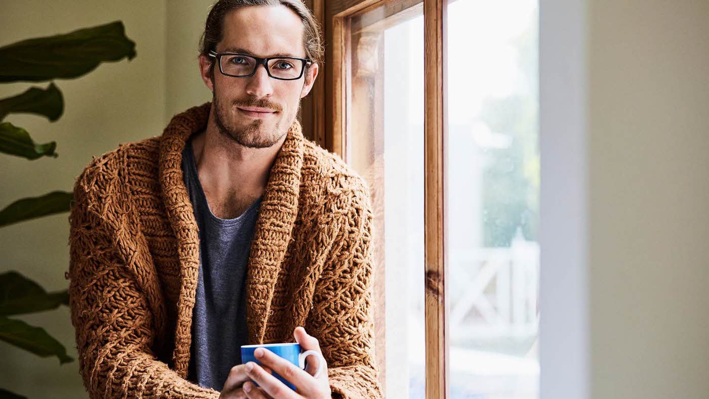 Mann med briller og brun strikkejakke som holder en kopp og sitter ved et vindu og ser i kamera