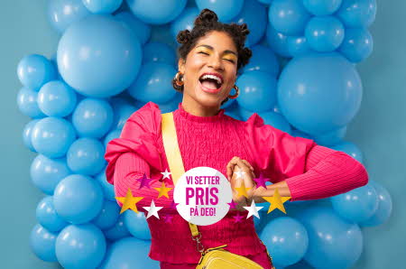 Smilende og takknenmlig kvinne foran blå ballongvegg med logoen "Vi setter PRIS på deg"