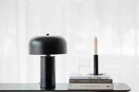 En svart lampe ved siden av tre bøker, en lysestake og et glass med vann på en svart hylle