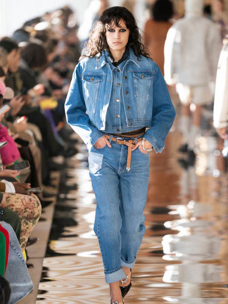 Damemodel på catwalk kledd i dongerijakke, skjorte og jeans, med belte i livet og pumps.
