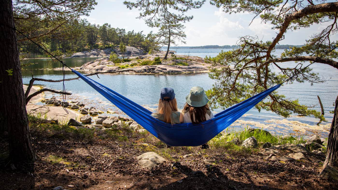 Sovepose i hengekøye Piknik ved sjøen To jenter sitter i hengekøye ved sjøen