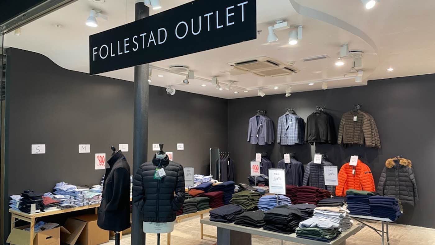 Follestad outlet butikk med hvitt tak, sorte veger og lyst gulv og salgsplakater på bord og over veggstativ