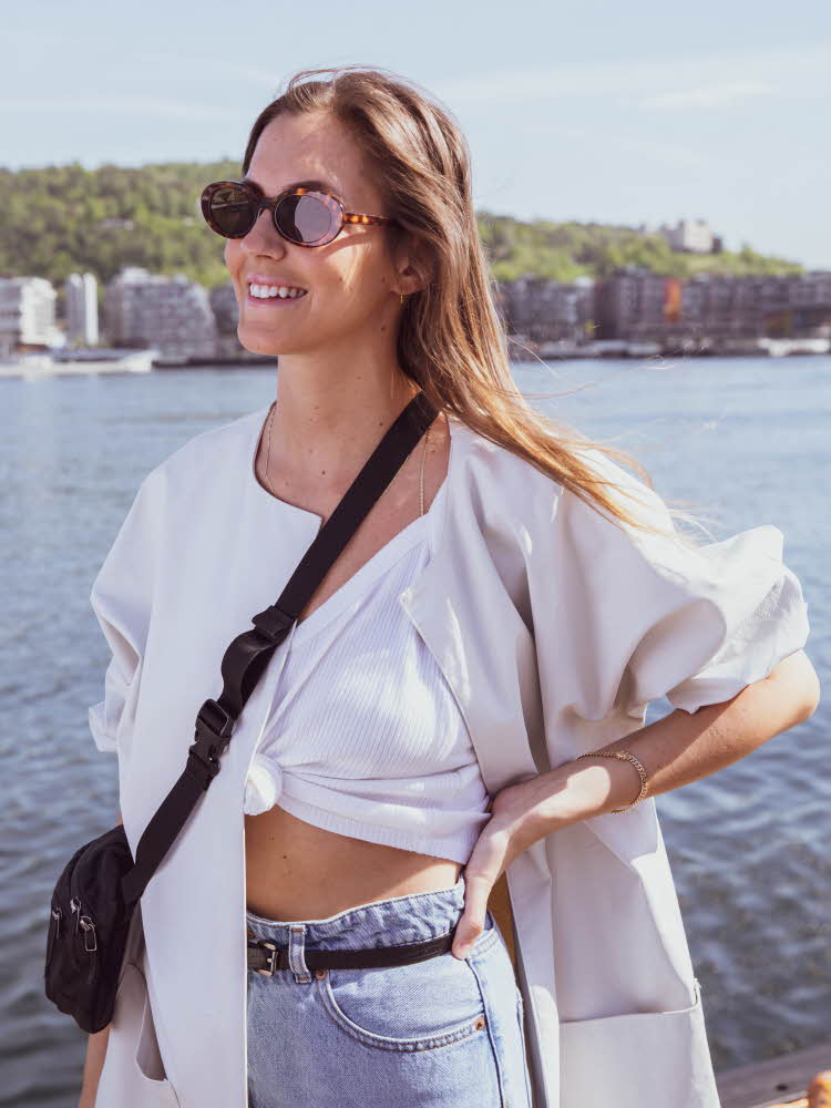 Brunette står på brygga iført lyse jeans, sort smalt belte, hvit tskjorte som er knytet og lys jakke