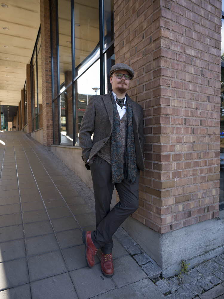 Mann i sixpence, brun rutete blazer og vest, hvit skjorte, tørkle, skjerf, grå bukse og brune sko.