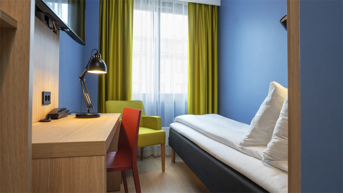 Garderobe, skrivebord, TV, blå vegger, gul stol, gule gardiner og enkeltseng på lite enkeltrom på Thon Hotel Astoria