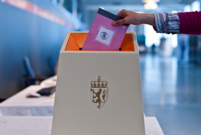 En hånd som holder en rosa stemmeseddel for stortingsvalget halvveis nedi en valgurne