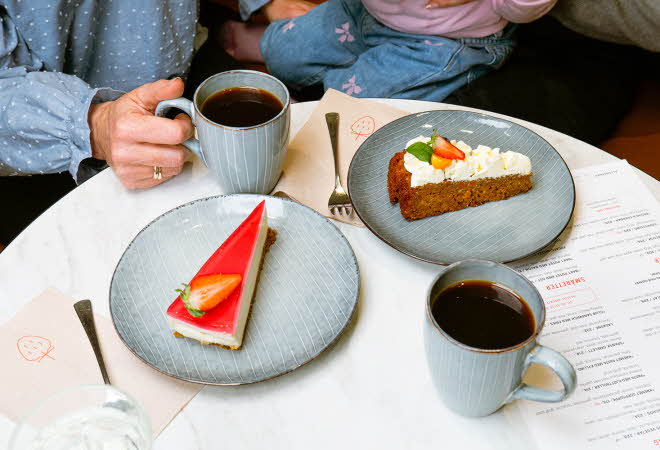 Et bord med to asjetter med kakebiter og to kopper kaffe, hvor en hånd holder den ene koppen