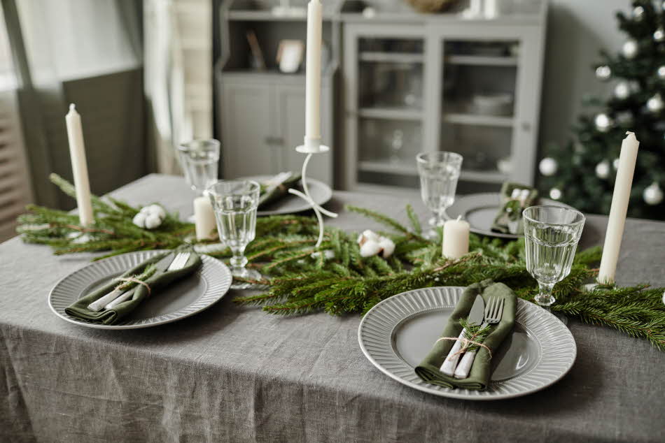 Matbord med grå linduk, pyntet med granbar og stearinlys. Fire fat, bestikk og glass. Juletre og vitrineskap i bakgrunnen.