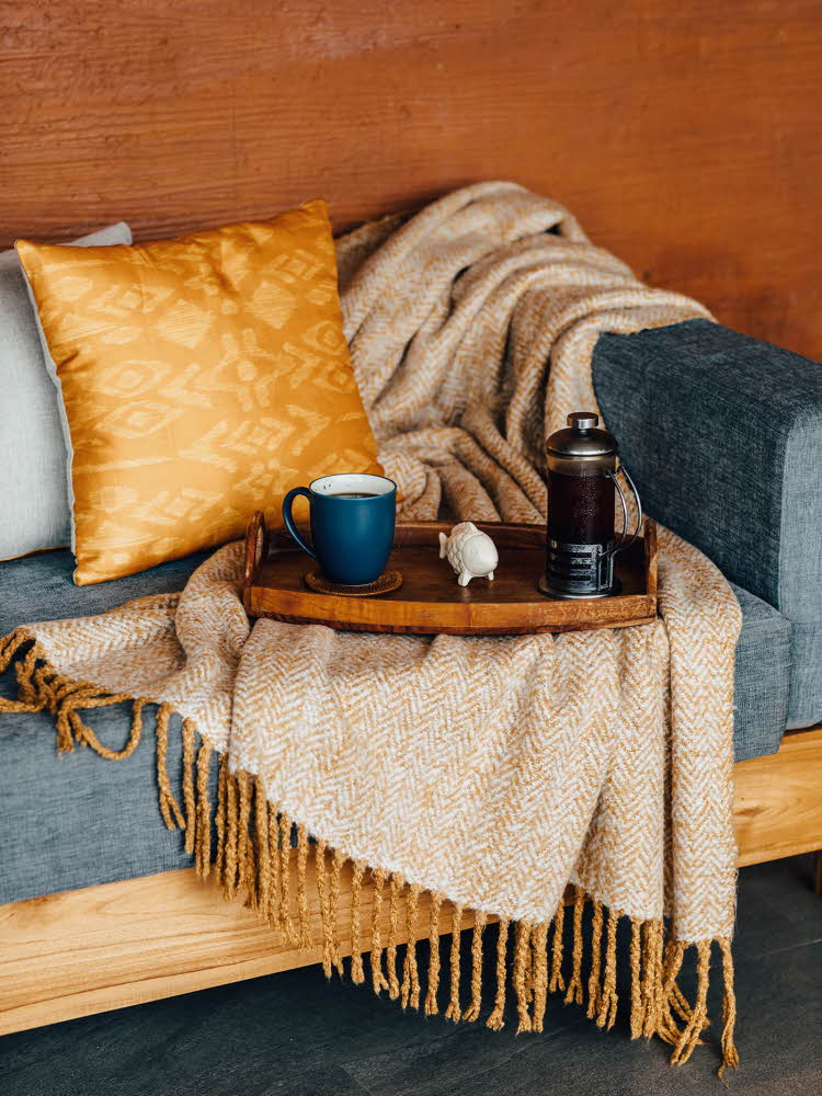Trebrett med kaffekopp og presskanne står oppå gult ullpledd i blå sofa med gul pute