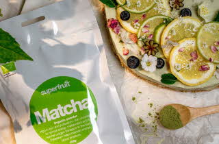 En pose med Superfruit Matcha ligger på et bord med sitron og lime og en skje med matcha pulver