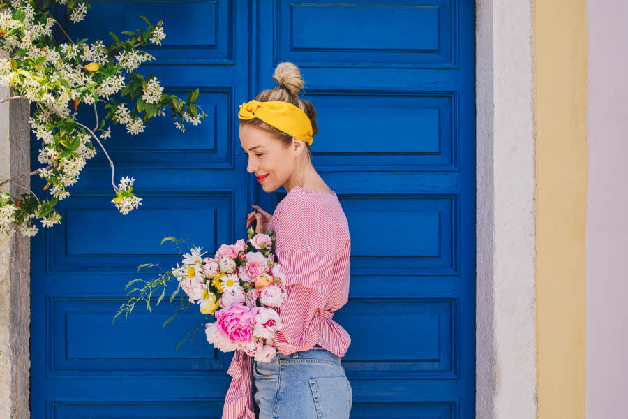 Dame iført gult hårbånd, rosa topp og lyse jeans står ved blå inngangsdør og holder en bukett blomster