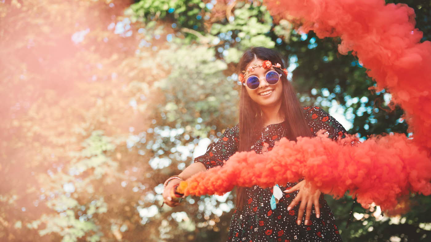 Jente med blomsterkrans og runde solbriller holder en rød/oransje fargebombe