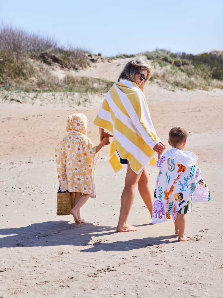 Dame med to barn som går på stranden med håndklær rundt seg
