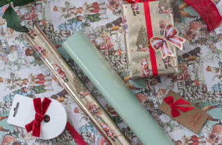 Gavepapir med julemotiv, ruller, bånd og innpakket gave med rødt sløyfebånd.