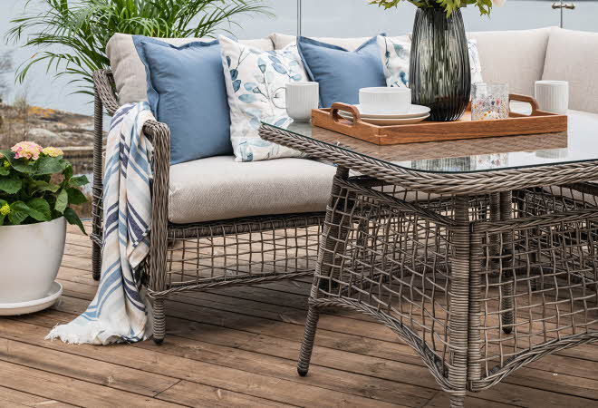 Uteområde med flettet sofa og bord, dekorert med puter og blomster.