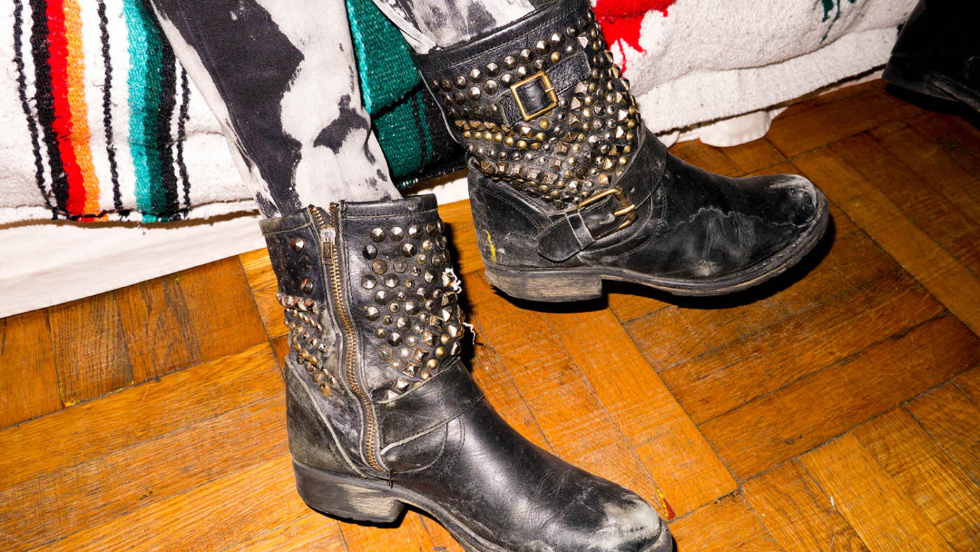 Nærbilde av sorte skinnboots med gullnagler og metallspenne med tregulv og sofa i bakgrunnen.