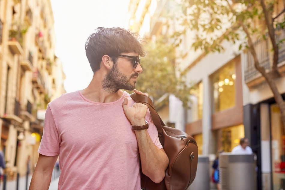 Mann med rosa t-skjorte og brun skinnbag over skulderen poserer på gate utendørs.
