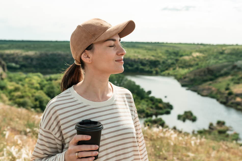 Dame med hvit- og blåstripete genser, mørk beige caps og kaffekopp i hånden nyter solen foran skog- og vannlandskap