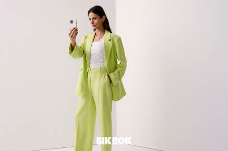 dame med grønn blazer, hvit topp, grønn dressbukse som poserer med en telefon i hånden