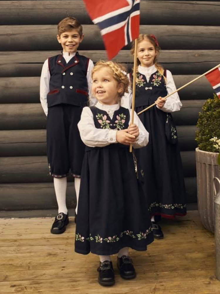 Tre barn med festdrakter, to jenter og en gutt. Jentene holder norske flagg.