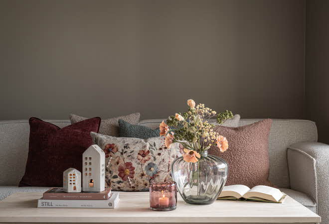 Stue med lys grå sofa, pynteputer og et sofabord med lyshus, bøker, blomster og et tent lys.