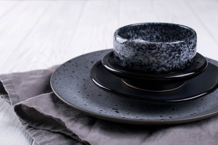 Skål og tre fat i mørke grå- og svarttoner med sjatteringer ligger oppå gråt håndkle på hvitmalt treflate