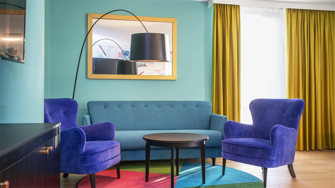 Turkis tapet, speil med gull ramme, gule gardiner, sittegruppe med turkis sofa, blå stoler og lite mørkt bord på fargerik teppe i business handicap room på Thon Hotel Storo i Oslo. 