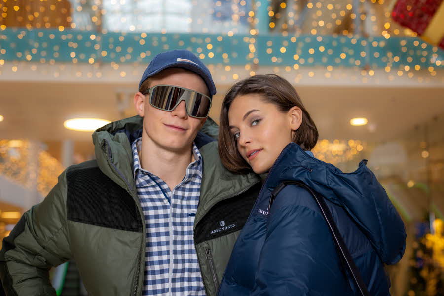 Ung mann og dame står inne på et kjøpesenter, han med caps, solbriller, dunjakke og skjort og hun med dunjakke.