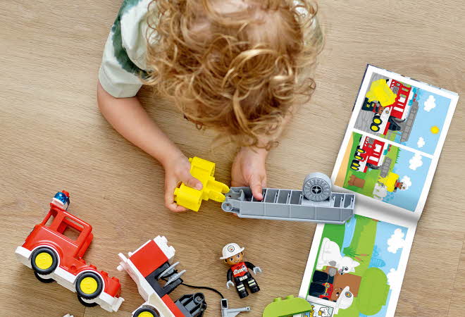 Barn bygger en LEGO Duplo brannbil på gulvet med instruksjonshefte ved siden av.