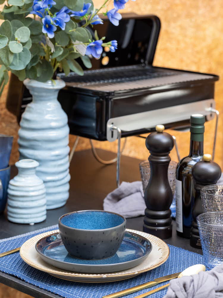 Detaljbilde av dekket bord med blått service, blomstervase med blomster og grill i bakgrunnen