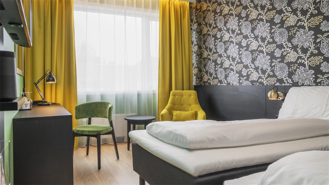 Separate senger, mørk mønstrete tapet med blomster, gule gardiner, gul stol med rundt bord og skrivebord med grønn stol i twin rom på Thon Hotel Kirkenes