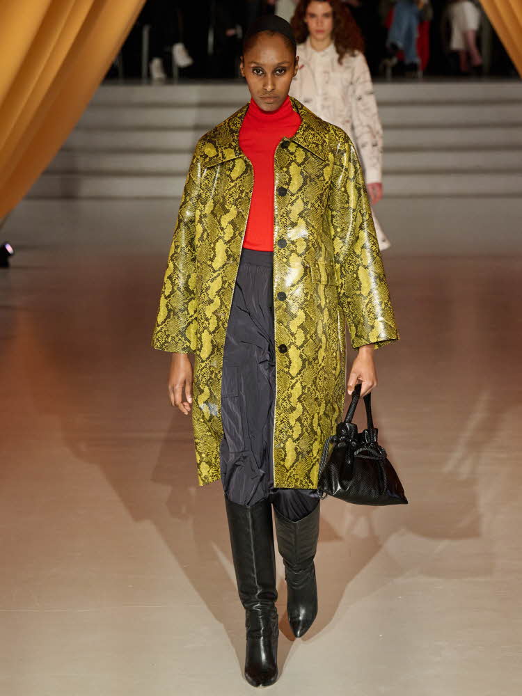 Damemodell på catwalk i sorte støvletter, grått skjørt, rød genser og gul frakk med slangeskinnmønster.