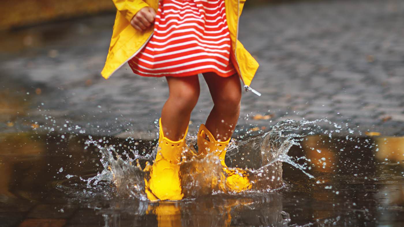 jente med gummistøvler og regnfrakk som hopper i en vanndam