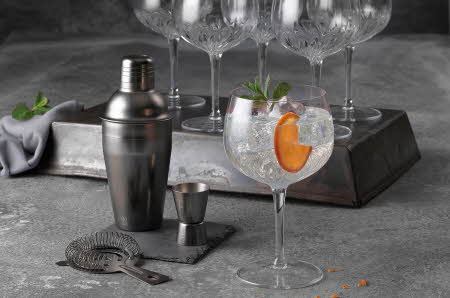 Et cocktailglass med is, appelsinskive og mynte står i sentrum, omgitt av shaker, målebeger og tomme krystallglass.