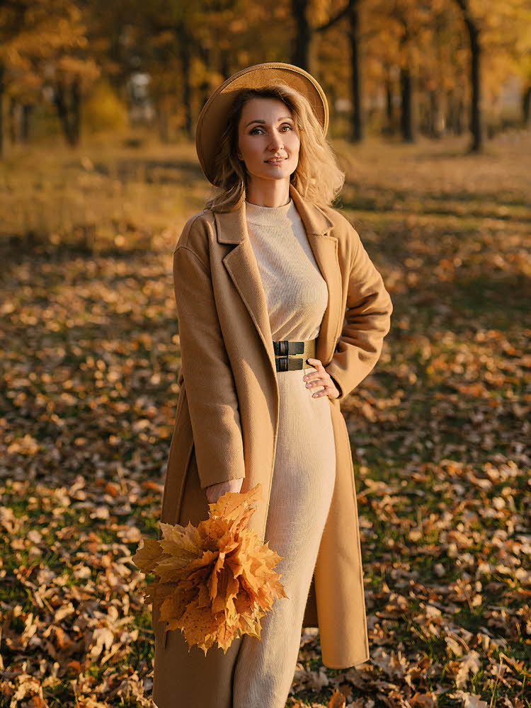 Dame med brun kjole og brun kåpe med mørkebrunt belte i midjen poserer med gule blader utendørs