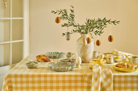 Et bord dekket til påskefrokost, med gul duk og en blomst med eggdekorasjoner på