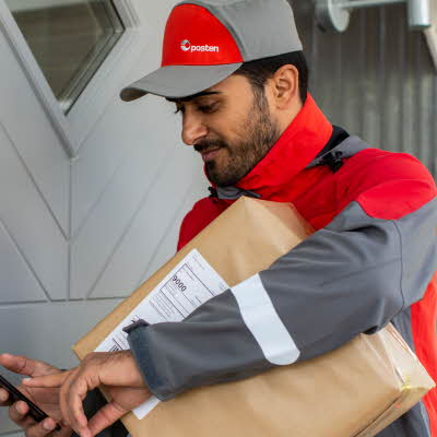 Mann iført arbeidsklær fra Posten står utenfor en dør med en pakke under armen som skal leveres